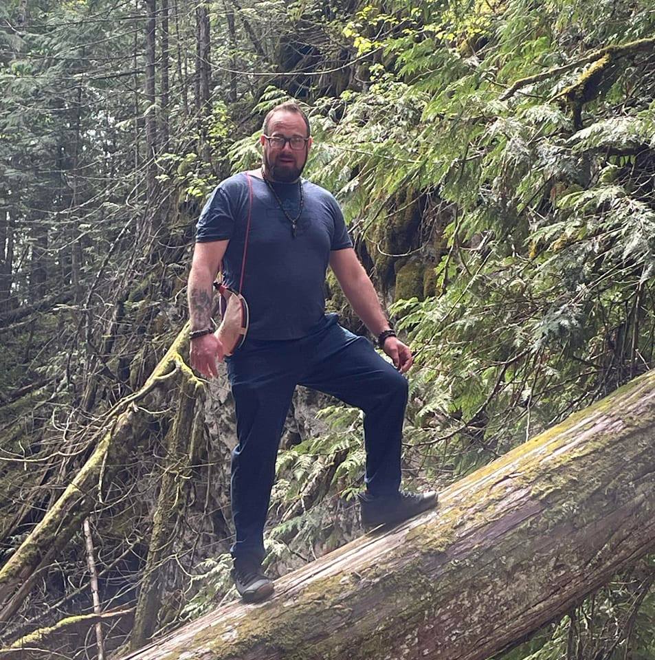 Bryan Sarge standing confidently on a moss-covered cedar tree in the forests of Shuswap, British Columbia — blending creativity, strength, and nature.